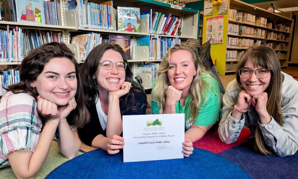 Photo of four women laying on the ground and posing with a piece of paper, in front of a bookshelf.